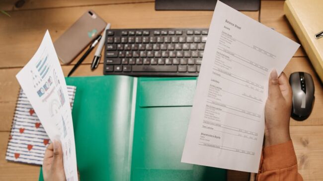 Hands holding and reviewing balance sheets over a desk with a keyboard and stationery. - Photo by Pavel Danilyuk on Pexels