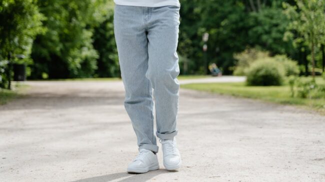 A close-up view of a person walking on a park pathway wearing blue jeans and white sneakers. - Photo by MART PRODUCTION on Pexels