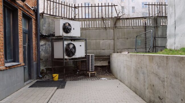 Air conditioning system located outside concrete shabby fence with metal barrier near industrial building from bricks and glass door - Photo by ready made on Pexels