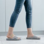 Close-up of a person wearing jeans and gray slippers, walking indoors. - Photo by MART PRODUCTION on Pexels